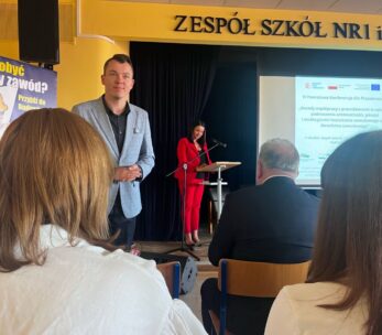 Mężczyzna w jasnoniebieskiej marynarce stoi przodem do kamery w klasie lub audytorium. Za nim na podium przemawia kobieta w czerwonym garniturze. Kilka osób siedzi, patrząc na ekran projekcyjny. Powyżej widnieje napis ZESPÓŁ SZKÓŁ NR1 im.