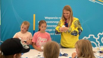 Kobieta w żółtej koszuli z niebieskimi wzorami prowadzi czwórkę dzieci wykonujących rękodzieło przy stole. Dzieci skupiają się na swoich projektach, trzymając białe przedmioty. Za nimi znajduje się turkusowy baner z logo i polskim tekstem. Na stole leżą nożyczki i przybory plastyczne.