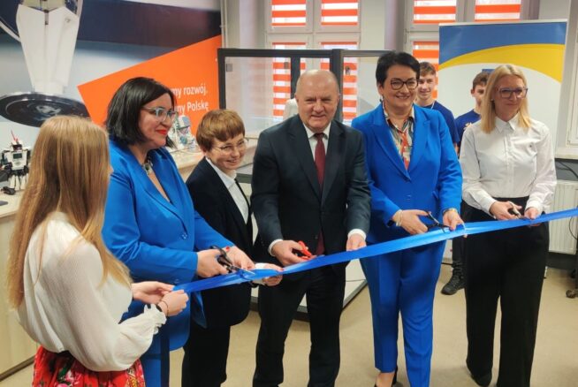 Grupa ludzi, w tym trzy kobiety w niebieskich garniturach i mężczyzna w ciemnym garniturze, przecina uroczystą niebieską wstęgę w pomieszczeniu. Dwie kobiety stoją po obu stronach, jedna w białej bluzce, a druga w białej koszuli z kolorową spódnicą. Tło stanowią plakaty i duże okna.