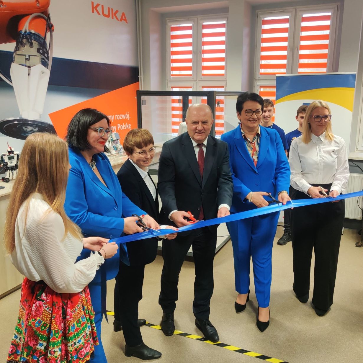 Grupa ludzi, w tym trzy kobiety w niebieskich garniturach i mężczyzna w ciemnym garniturze, przecina uroczystą niebieską wstęgę w pomieszczeniu. Dwie kobiety stoją po obu stronach, jedna w białej bluzce, a druga w białej koszuli z kolorową spódnicą. Tło stanowią plakaty i duże okna.