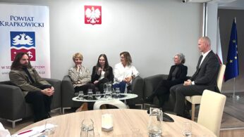 Sześć osób siedzi w półokręgu na szarych krzesłach w formalnej sali konferencyjnej. Jedna kobieta, trzymając mikrofon, mówi, podczas gdy inni słuchają. Za nimi znajdują się flagi Polski i UE oraz baner Powiatu Krapkowickiego. Na pierwszym planie okrągły stół z kieliszkami.