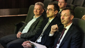 Czterech mężczyzn w strojach biznesowych siedzi w fotelach na widowni, uważnie obserwując coś poza kamerą. Jeden mężczyzna z przodu trzyma okulary i schowek, podczas gdy pozostali mają poważne miny. Otoczenie wydaje się formalne i profesjonalne.
