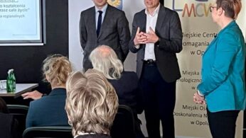 Trzy osoby stoją i przemawiają do siedzącej publiczności w sali konferencyjnej. Za nimi znajduje się slajd prezentacji i baner z logo MCWP. Słuchacze zwróceni są twarzami do przodu, a podłogę pokrywa wzorzysty dywan.
