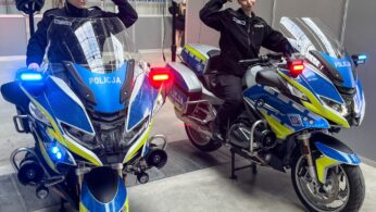 Dwie kobiety w czarnych policyjnych mundurach dumnie pozują obok niebiesko-żółtych motocykli POLICJA. Każda z kobiet zgina ramię w silnym geście; jedna stoi przy swoim motocyklu, druga siedzi okrakiem. Kaski spoczywają na motocyklach, a niebieskie światła są podświetlone.
