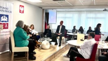 Grupa ludzi siedzi na platformie w nowoczesnej sali konferencyjnej, angażując się w dyskusję panelową. Inni przyglądają się ze stolików. Na ścianie wisi polska flaga, godło z białym orłem i sztandar Powiatu Krapkowickiego. Duże okna wpuszczają światło dzienne.
