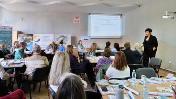 Grupa dorosłych siedzi przy stołach w sali konferencyjnej, naprzeciwko prezentera w czerni w pobliżu wyświetlanego ekranu. Na stołach leżą papiery, butelki i foldery. Uczestnicy uważnie słuchają, a na lewej ścianie widoczne są banery.