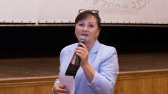 Kobieta w jasnoniebieskim garniturze trzyma mikrofon i kartkę papieru, przemawiając przed drewnianą sceną. Za nią znajduje się biały ekran z niewyraźnym tekstem i niewyraźnym rysunkiem uśmiechniętych twarzy. Wygląda na to, że wygłasza prezentację lub przemówienie.