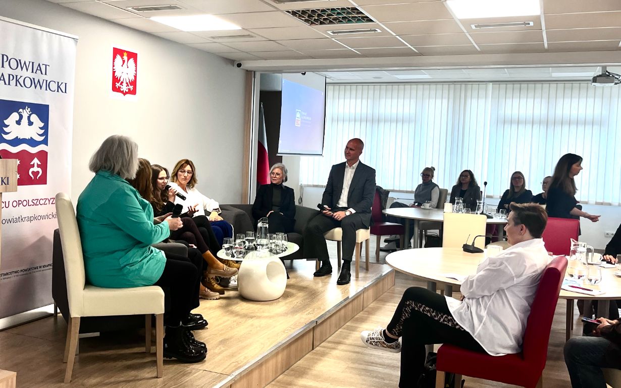Grupa ludzi siedzi na platformie w nowoczesnej sali konferencyjnej, angażując się w dyskusję panelową. Inni przyglądają się ze stolików. Na ścianie wisi polska flaga, godło z białym orłem i sztandar Powiatu Krapkowickiego. Duże okna wpuszczają światło dzienne.