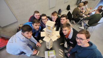 Ośmiu młodych ludzi gromadzi się wokół stołu, uśmiechając się do kamery. Na stole stoi wysoka drewniana wieża z klocków, którą wspólnie zbudowali. Kolejne grupy pracują nad podobnymi projektami w jasnym, nowoczesnym pomieszczeniu o szarych ścianach.