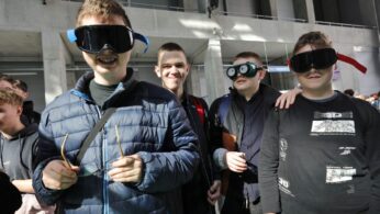 Grupa czterech uśmiechniętych młodych mężczyzn stoi w pomieszczeniu. Trzech nosi duże, zabawne gogle z przerysowanymi wzorami, a jeden trzyma okulary. Są otoczeni przez innych ludzi, a światło słoneczne wpada przez duże okna za nimi.