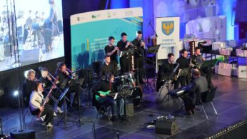 Na scenie występuje zespół jazzowy z instrumentami dętymi blaszanymi, drewnianymi, gitarą, fortepianem i perkusją. Muzycy siedzą z pulpitami na nuty. Za nimi znajdują się duże banery i ekran z projekcją na żywo. Scena jest ustawiona w nowoczesnym pomieszczeniu z widocznymi światłami i sprzętem.