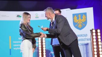 Mężczyzna w garniturze podaje rękę uśmiechniętej młodej kobiecie, wręczając jej nagrodę na scenie. Inny mężczyzna stoi za nimi, trzymając schowek. W tle widoczne są niebiesko-białe banery i żółty herb z orłem.