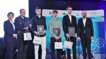 Sześć osób stoi w rzędzie na scenie, cztery trzymają certyfikaty i torby z prezentami. Są ubrani formalnie. Za nimi znajduje się niebieskie tło z logo i stylizowanymi kołami zębatymi. Scena wygląda na ceremonię wręczenia nagród lub wyróżnień.
