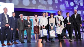 Grupa jedenastu osób, w tym zarówno dorosłych, jak i nastolatków, stoi na scenie pod jasnymi światłami. Niektórzy trzymają ozdobne torby z prezentami. Są ubrani formalnie, pozują i uśmiechają się do zdjęcia przed banerami i wyświetlanym wzorzystym tłem.