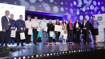 Grupa trzynastu osób stoi na oświetlonej scenie, trzymając certyfikaty i torby z prezentami. Ubrani są w stroje biznesowe lub eleganckie. Niebieskie i fioletowe światła tworzą okrągłe wzory na ścianie za nimi. Po prawej stronie widoczny jest baner z żółto-niebieskim herbem.