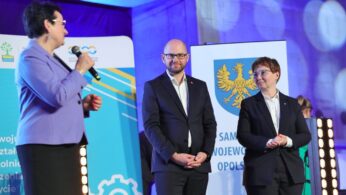 Trzy osoby w oficjalnych strojach stoją na scenie z mikrofonami; kobieta po lewej stronie przemawia, podczas gdy dwoje innych, mężczyzna i kobieta, słuchają i uśmiechają się. W tle widoczne są kolorowe banery z polskim tekstem i herbem. Jasne światła sceniczne są z przodu.
