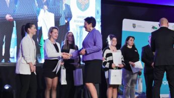 Kobieta w fioletowej marynarce podaje rękę młodej kobiecie w czerni na scenie. Inne kobiety stoją w kolejce, trzymając certyfikaty i torby z prezentami. Obecni są ludzie w garniturach. W tle widoczny jest duży ekran i banery z logo.
