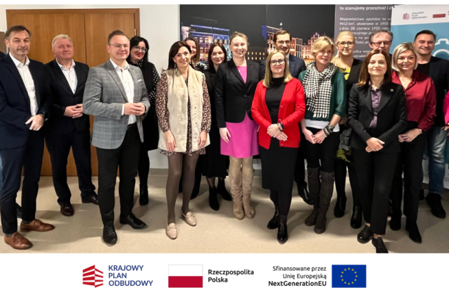 Grupa 16 profesjonalnie ubranych osób stoi w pomieszczeniu, uśmiechając się do kamery. Za nimi znajdują się plakaty z tekstem i obrazami. Pod zdjęciem widnieją trzy logotypy i nazwy - Krajowy Plan Odbudowy, Rzeczpospolita Polska i Unia Europejska.
