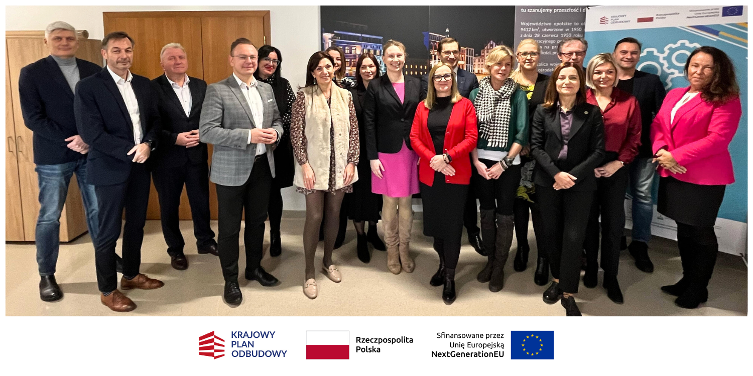 Grupa 16 profesjonalnie ubranych osób stoi w pomieszczeniu, uśmiechając się do kamery. Za nimi znajdują się plakaty z tekstem i obrazami. Pod zdjęciem widnieją trzy logotypy i nazwy - Krajowy Plan Odbudowy, Rzeczpospolita Polska i Unia Europejska.