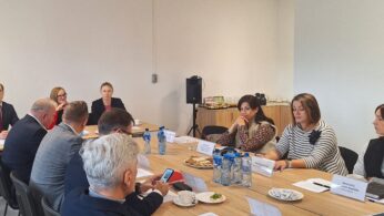 Grupa ludzi, zarówno mężczyzn, jak i kobiet, siedzi wokół prostokątnych stołów w sali konferencyjnej. Niektórzy rozmawiają, inni słuchają. Na stołach leżą papiery, woda butelkowana i przekąski. W tle na tle prostej ściany widoczny jest głośnik i bufet.