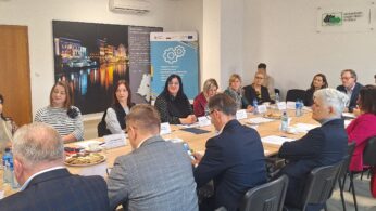 Grupa czternastu osób, mężczyzn i kobiet, siedzi przy stole konferencyjnym z papierami, butelkami wody i przekąskami. Wydają się być zaangażowani w dyskusję. Za nimi znajdują się banery i znaki na ścianach, z których jeden przedstawia budynki miejskie, a drugi logo.