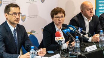 Trzy osoby w strojach biznesowych siedzą przy stole konferencyjnym z mikrofonami i butelkami wody. Kobieta w centrum przemawia, podczas gdy dwaj mężczyźni słuchają. Za nimi znajdują się banery z logo i polskim tekstem, wskazujące na oficjalne wydarzenie lub konferencję prasową.
