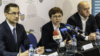 Trzy osoby w strojach biznesowych siedzą przy stole z mikrofonami i butelkami wody, przemawiając na konferencji prasowej. Za nimi znajdują się banery z polskim tekstem i logo władz regionalnych. Kobieta w centrum mówi do mikrofonów, otoczona przez dwóch mężczyzn.