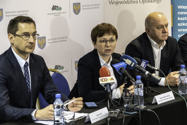 Trzy osoby w strojach biznesowych siedzą przy stole z mikrofonami i butelkami wody, przemawiając na konferencji prasowej. Za nimi znajdują się banery z polskim tekstem i logo władz regionalnych. Kobieta w centrum mówi do mikrofonów, otoczona przez dwóch mężczyzn.