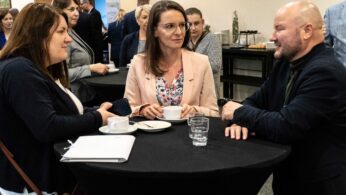 Trzy osoby stoją wokół czarnego okrągłego stołu na profesjonalnym wydarzeniu, rozmawiając i trzymając filiżanki z kawą. Dwie kobiety i jeden mężczyzna są zaangażowani w rozmowę. Inni uczestnicy i przekąski są w tle. Ustawienie wydaje się formalne i towarzyskie.
