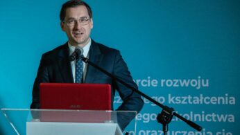 Mężczyzna w garniturze i okularach przemawia na podium z mikrofonem. Na podium stoi czerwony laptop ASUS. Tło jest turkusowe z białym, częściowo widocznym polskim tekstem. Oświetlenie podkreśla twarz i garnitur mówcy.