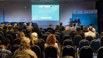 Sala konferencyjna z uczestnikami siedzącymi w rzędach naprzeciwko sceny. Mężczyzna stoi na podium, prowadząc prezentację obok ekranu wyświetlającego slajdy z tekstem i grafiką sprzętową. W pokoju znajdują się niebieskie krzesła i banery na ścianach.