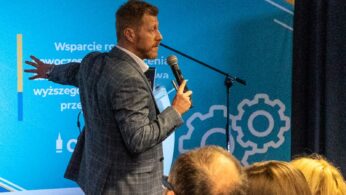 Mężczyzna w kraciastej marynarce mówi do mikrofonu, gestykulując ręką podczas prezentacji. Stoi przed niebieskim i turkusowym tłem z tekstem i grafiką, zwrócony w stronę publiczności siedzącej na pierwszym planie.