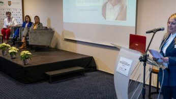 Kobieta w niebieskim garniturze przemawia na podium z mikrofonem i laptopem, podczas gdy cztery osoby siedzą na panelu scenicznym obok. Za nimi wyświetlany jest slajd prezentacji. Wygląda to na formalną konferencję lub seminarium.