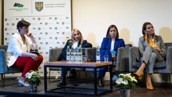 Cztery kobiety siedzą na scenie przed białym tłem z polskimi logo. Dwie kobiety noszą niebieskie garnitury, jedna szary garnitur, a jedna białą kurtkę z czerwonymi spodniami. Butelki z wodą i szklanki stoją na stoliku do kawy. Na podłodze obok stoją białe kwiaty doniczkowe.