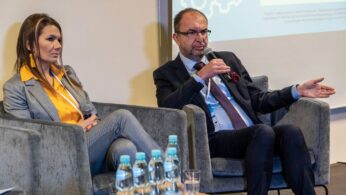 Kobieta i mężczyzna w strojach biznesowych siedzą na szarych fotelach, przemawiając podczas panelu. Kobieta trzyma mikrofon, a mężczyzna, gestykulując jedną ręką, również przemawia. Na stole przed nimi stoją butelki wody. Za nimi wyświetlana jest prezentacja.