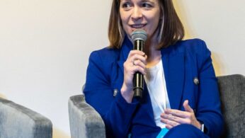 Kobieta o prostych, sięgających ramion brązowych włosach ma na sobie jasnoniebieski garnitur i białą bluzkę. Siedzi na szarym krześle, trzyma mikrofon i mówi, a drugą ręką gestykuluje. Tło jest gładkie i jasne.