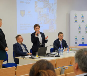 Cztery osoby stoją z przodu klasy lub sali konferencyjnej. Jedna kobieta stoi, trzech mężczyzn siedzi, a jeden z siedzących mężczyzn mówi do mikrofonu. Na ścianie za nimi wyświetlana jest prezentacja, a na stole leżą butelki z wodą i laptopy.