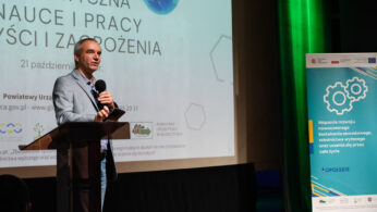 Mężczyzna w garniturze przemawia do mikrofonu na scenie podczas konferencji. Za nim znajduje się ekran projekcyjny z polskim tekstem i niebiesko-zielony baner z kołami zębatymi i ikonami, promujący nowoczesne kształcenie i szkolenie zawodowe.