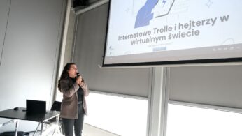 Osoba stoi trzymając mikrofon i mówi przed ekranem projektora. Na ekranie wyświetlany jest slajd w języku polskim z ilustracją osoby przy komputerze i tekstem: 