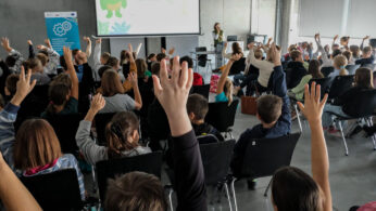 Klasa wypełniona dziećmi siedzącymi w rzędach, wiele z nich podnosi ręce. Z przodu kobieta przemawia w pobliżu ekranu projektora wyświetlającego zieloną postać z kreskówki i tekst. Pomieszczenie ma nowoczesny, industrialny wystrój z betonowymi ścianami i dużymi oknami.