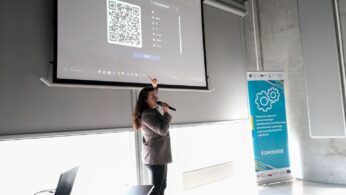 Kobieta stoi przed dużym ekranem projekcyjnym, wskazując na wyświetlany na nim kod QR. Trzyma mikrofon i wydaje się prowadzić prezentację. Na stole znajduje się laptop, a po prawej stronie pionowy baner z kołami zębatymi i tekstem.