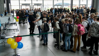 Duża grupa dzieci z plecakami stoi w kolejce wewnątrz nowoczesnego budynku. Dorośli prowadzą je w kierunku stanowiska rejestracyjnego ozdobionego kolorowymi balonami. W tle widać więcej ludzi i stoisk z dużymi oknami wpuszczającymi światło dzienne.