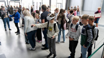 Grupa dzieci stoi w kolejce, podczas gdy chłopiec z przodu korzysta z zestawu VR i kontrolerów. Dorośli i inne dzieci obserwują. Scena jest wewnątrz, jasna z dużymi oknami, a w tle są banery i ludzie.