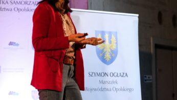Kobieta z długimi ciemnymi włosami, ubrana w czerwoną marynarkę, koszulę w paski i dżinsy, mówi do mikrofonu trzymając w ręku pilota. Stoi przed banerem z polskim godłem i tekstem wspominającym Szymona Ogłazę i województwo opolskie.