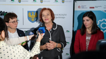 Trzy kobiety stoją przed banerami podczas wywiadu. Kobieta w środku mówi i uśmiecha się, trzymając ręce razem. Reporterzy trzymają w jej kierunku mikrofony i urządzenia nagrywające. Dwie inne kobiety stoją po obu stronach, jedna uważnie, a druga lekko odwrócona.