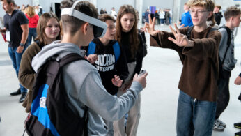 Grupa nastolatków stoi w pomieszczeniu. Jeden z nich nosi gogle VR i trzyma kontroler, podczas gdy inny gestykuluje animacyjnie uniesionymi rękami. Inni przyglądają się z zainteresowaniem, nosząc zwykłe ubrania i plecaki. W tle widać więcej osób.