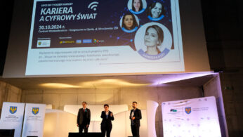 Trzy osoby w strojach biznesowych stoją na scenie, przemawiając do publiczności. Nad nimi znajduje się duży ekran wyświetlający cyfrowy plakat wydarzenia ze zdjęciami czterech kobiet, szczegółami wydarzenia i logo. Miejsce wydarzenia charakteryzuje się nowoczesnym designem i dwoma stojącymi banerami.
