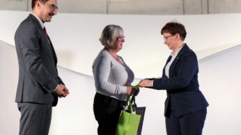 Trzy dorosłe osoby stoją w pomieszczeniu. Kobieta w garniturze wręcza kartkę lub papier drugiej kobiecie trzymającej zielono-czarną torbę. Mężczyzna w szarym garniturze i czerwonym krawacie stoi po lewej stronie, obserwując wymianę. Tło jest gładkie i białe.