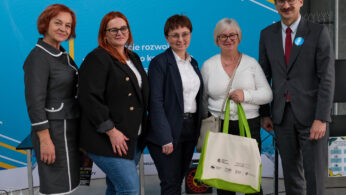 Pięć dorosłych osób stoi razem w pomieszczeniu, uśmiechając się do kamery. Cztery kobiety i jeden mężczyzna ubrani są w strój biznesowy lub elegancki. Jedna z kobiet trzyma kremową torbę z zielonymi uchwytami. W tle znajduje się niebiesko-biały wyświetlacz wydarzenia.