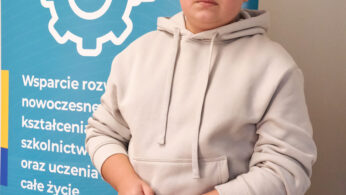 Młody chłopak w szarej bluzie z kapturem trzyma beżową torbę na ramię, stojąc przed niebieskim banerem Krajowego Planu Odbudowy finansowanym ze środków UE, na którym widnieją ikony sprzętu i logo województwa opolskiego.
